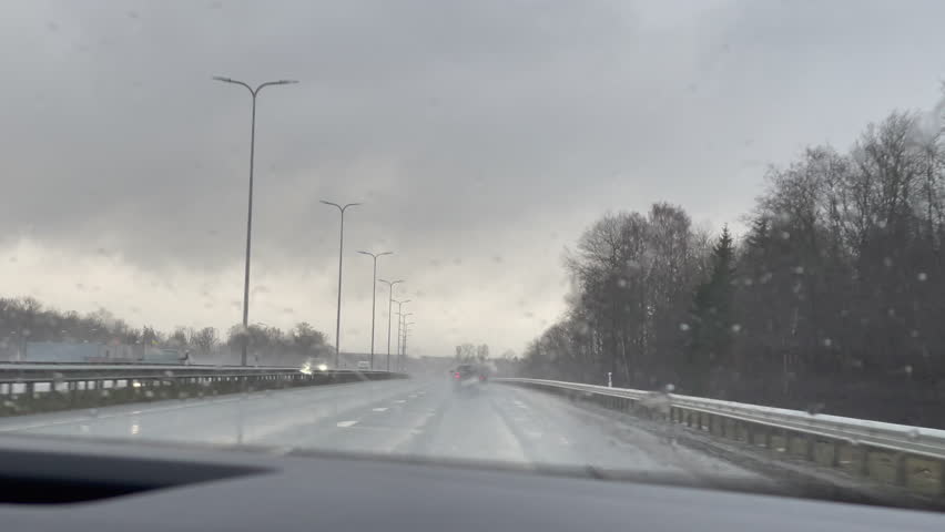 Car front window view driving on a rainy stormy highway with cloudy low light winter afternoon foggy misty weather raining cars passing and trees without leaves next to the road in Estonia, icy roads  Royalty-Free Stock Footage #1099536631