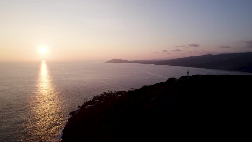Cinematic aerial of sun setting over the water off the coast of Oahu, Hawaii. Mountain ranges surround Honolulu in the Maunalua Bay area