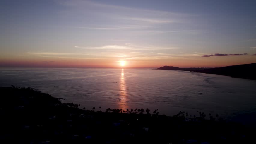 Cinematic aerial of sun setting over the water off the coast of Oahu, Hawaii. Mountain ranges surround Honolulu in the Maunalua Bay area