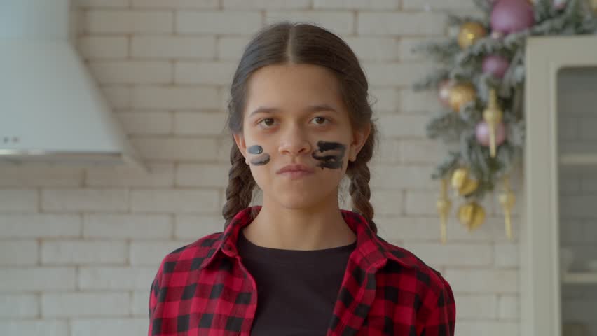 Confident attractive Hispanic adolescent girl with pigtails wearing eye black under eyes, showing watching you gesture, expressing determination and readiness indoors.