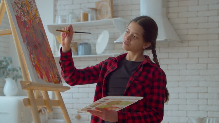 Engrossed in creative process talented adorable Latin teen girl with pigtails expressing active imagination , inspiration and ideas, painting picture on easel using paintbrush and colorful palette. | Shutterstock HD Video #1099539231