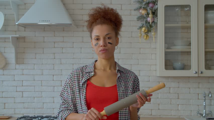 Portrait of strict determined attractive African American female wearing eye black, holding rolling pin and making aggressive gesture, showing irritation , fury and anger indoors.