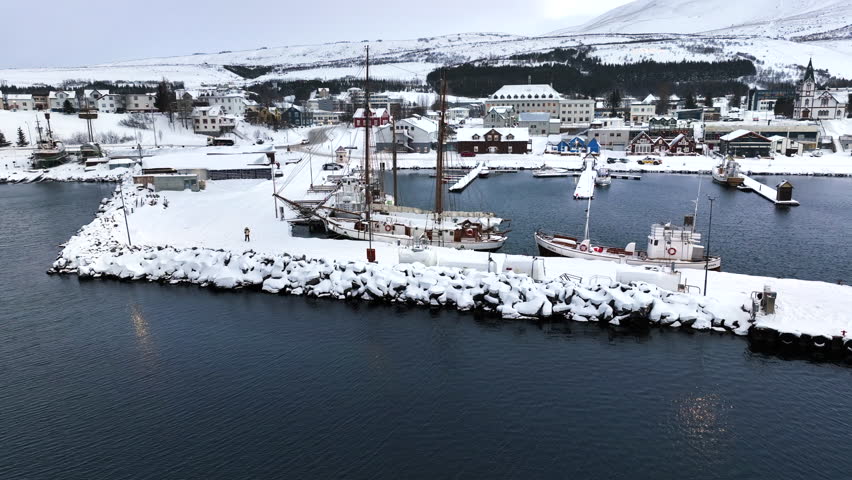 Large sailboat is moored in the harbor of Husavic while the harbor is covered with white snow. Drone panning shot
