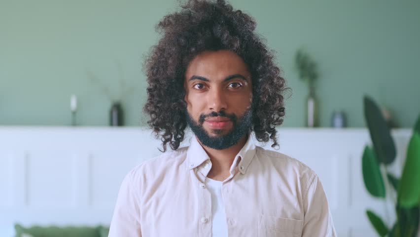 Young happy curly Arabian man millennial looking at camera with snow-white smile wishing to share positive emotions and good mood dressed in casual style stands in spacious apartment | Shutterstock HD Video #1099547385