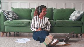 Young calm focused African American woman engage in self-education by watching video lessons and training webinars on Internet in laptop and taking notes sits on floor in living room near sofa
