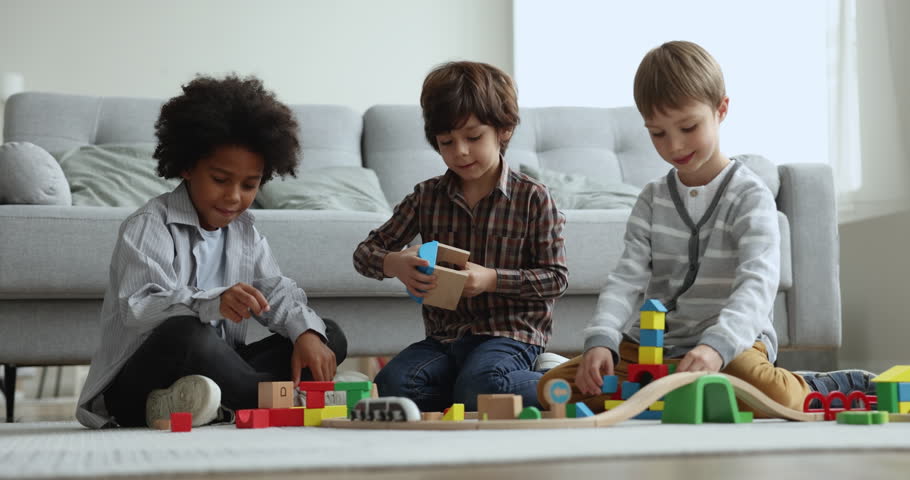 Three little multiethnic boys, best friends gathered in cozy living room play wooden railroad, enjoy communication using playthings. Friendship, cousins, family spend time at home, playtime, childhood