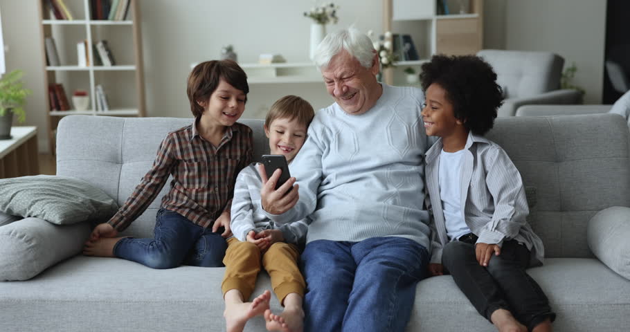 Elderly 80s grandad spend leisure with three little multi ethnic grandchildren, sit on sofa having fun using new mobile application, making video call, watching content on internet. Modern tech usage | Shutterstock HD Video #1099548829