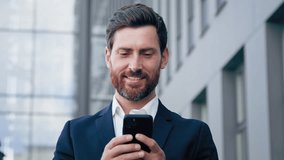 Close-up happy handsome bearded caucasian man looking at phone screen browsing social network using wifi successful businessman chatting online on smartphone smiling looks at camera posing outdoors