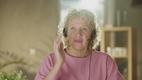 Mature adult curly haired blonde woman sitting at table, looking at camera, listening and speaking. Online remote communication concept