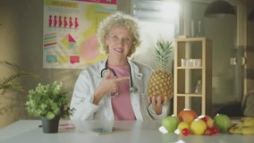 A mature female doctor or nutritionist sitting at the table with different fruits and pointing at pineapple - Powered by Shutterstock - Get 15% off with code: PIKWIZARD15