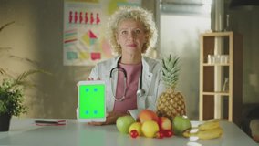 Mature female doctor or nutritionist sitting at the table, holding smart tablet with different fruits and vegetables - Powered by Shutterstock - Get 15% off with code: PIKWIZARD15
