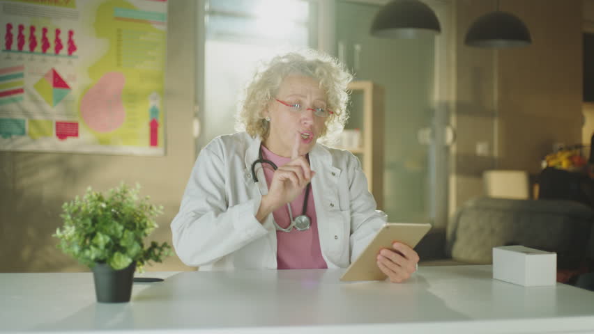 Mature female doctor sitting at desk having video call with patient using her digital tablet | Shutterstock HD Video #1099551381