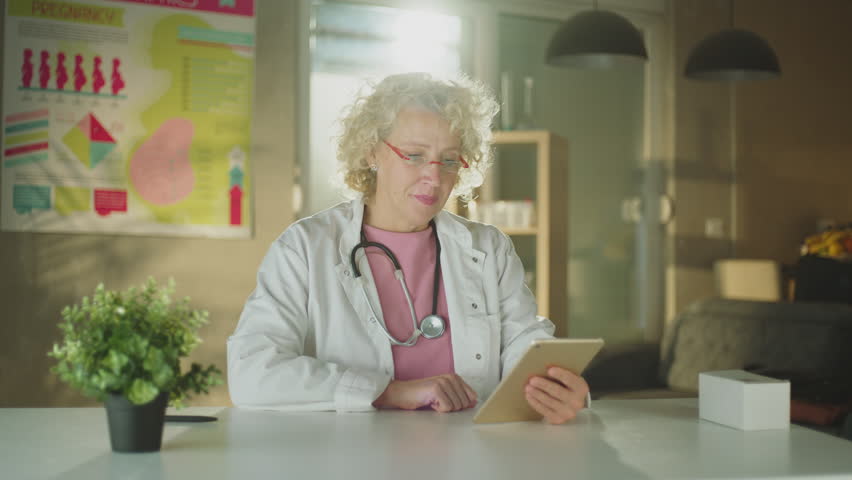 Mature female doctor sitting at desk having video call with patient using her digital tablet | Shutterstock HD Video #1099551383
