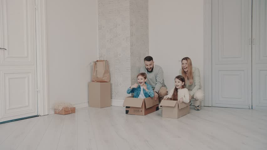 Mom and Dad are pushing cardboard boxes with cute little kids. Daughter and son are playing with their parents, having fun on the day of moving to a new apartment. New house. Happy family. | Shutterstock HD Video #1099551929