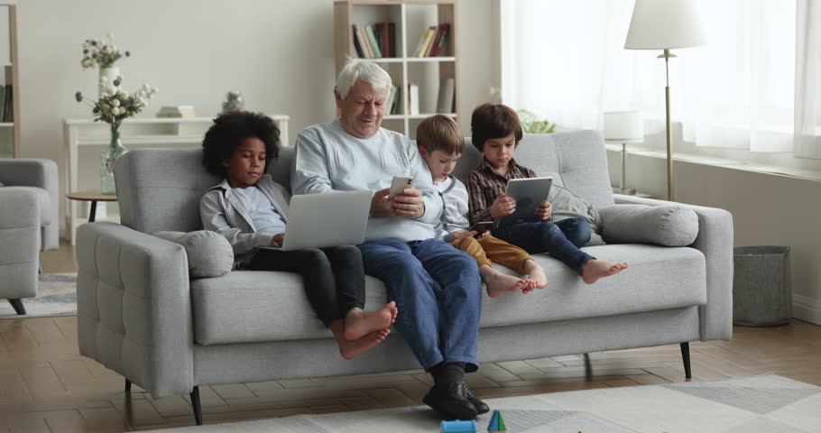 Older great-grandad with little grandkids use diverse electronic devices, sit on sofa with phones, digital tablet, laptop, focused in videogames, web surfing internet, play virtual games. Tech overuse | Shutterstock HD Video #1099552101