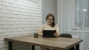 Overweight woman in glasses sits at wooden table in apartment and makes video call through tablet. Lady waves hand in greeting, smiles, listens to interlocutor, nods her head, medium shot.
