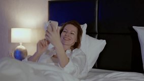 beautiful adult woman viewing funny video in internet by smartphone, lying in bed in evening