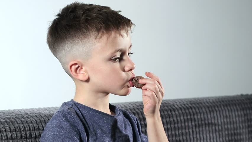 A little boy opening sweets and taking pastila his mouth while sitting on a cozy sofa. A child enjoys candy | Shutterstock HD Video #1099555863