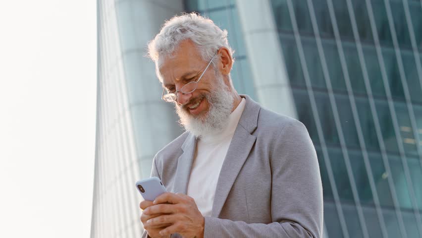 Happy older mature stylish professional business man, smiling senior 60 years old businessman wearing suit using smartphone using mobile cell phone tech standing outdoor in big city office district.