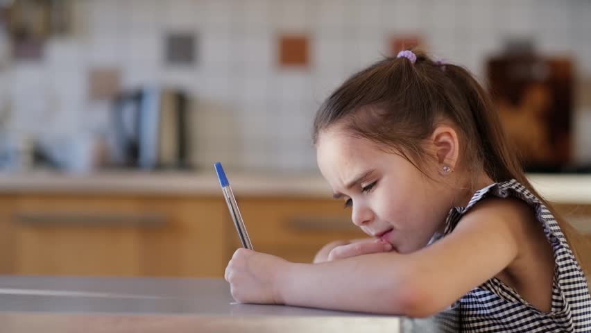 A girl of European appearance in a plaid dress is doing homework for the school at the table in the kitchen. The student thought about the example. The child draws with a pen on paper. High quality | Shutterstock HD Video #1099556569