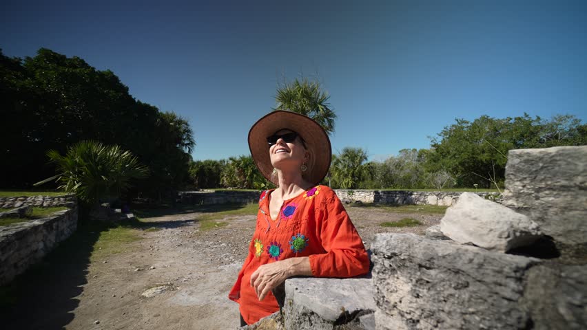 Camera orbits mature woman wearing ethnic clothes, sunglasses, hat taking in the stone ruins of Xcambo Mayan pyramid in Yucatan Mexico. Concept of adventure is ageless.