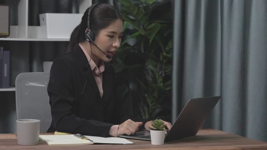 Businesswoman wearing headset and microphone working at her desk and using laptop. Enthusiastic female employee utilizing laptop for remote online meeting, business video conference in the office. | Shutterstock HD Video #1099560113