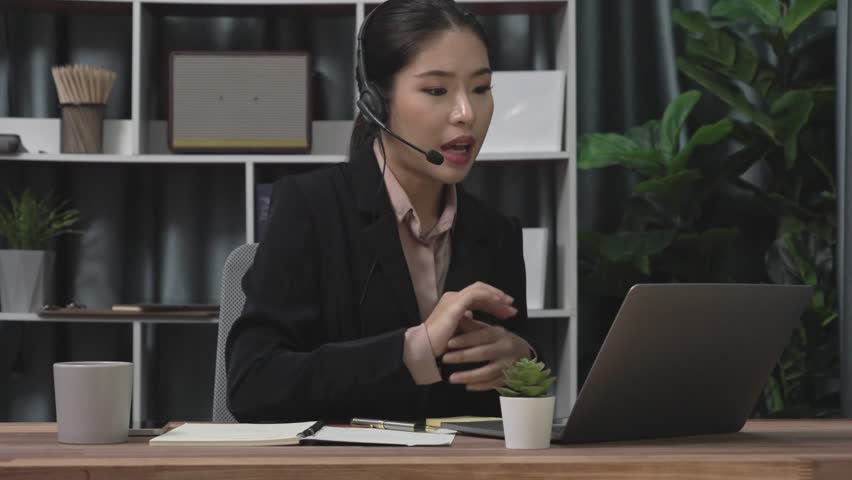Businesswoman wearing headset and microphone working at her desk and using laptop. Enthusiastic female employee utilizing laptop for remote online meeting, business video conference in the office. | Shutterstock HD Video #1099560179