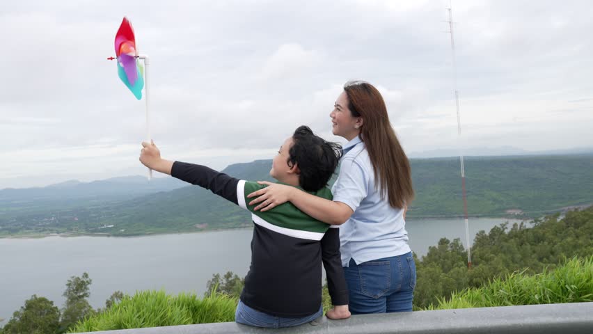 A progressive woman and her son are on vacation, enjoying the natural beauty of a lake at the bottom of a hill while the boy carries a toy windmill.