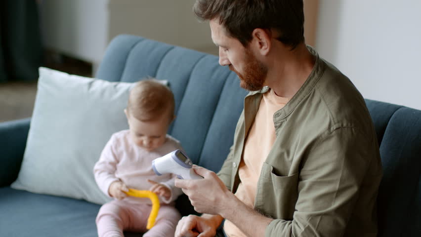 Kid sickness. Young caring father measuring body temperature of his playful baby daughter and seeing fever, feeling worried and surprised at home, tracking shot, slow motion, empty space | Shutterstock HD Video #1099560307