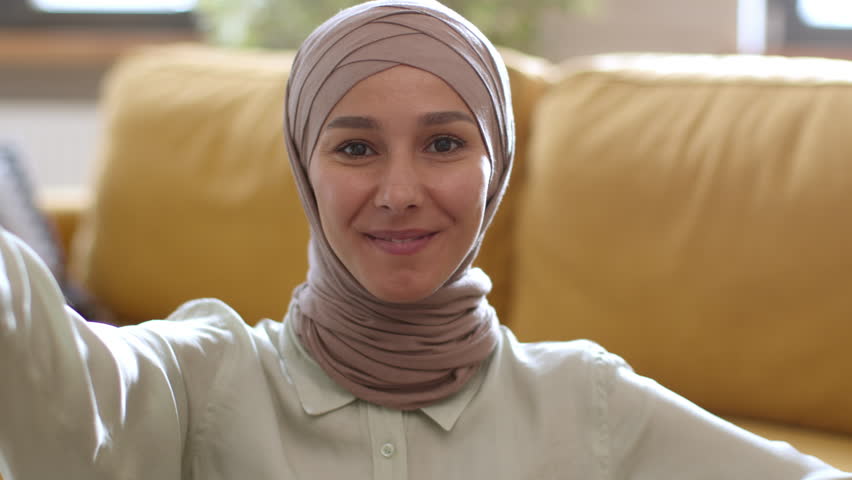 Video communication concept. Phone webcam pov portrait of young excited muslim woman in hijab video calling to friends, waving hand and telling great news at home Royalty-Free Stock Footage #1099560349