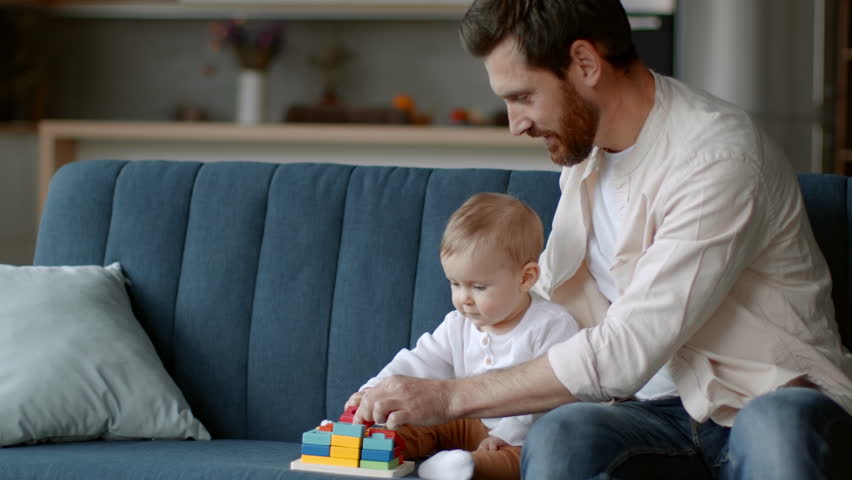 Early development. Caring young father playing with his cute baby daughter at home, searching new wooden toy on sofa, tracking shot, slow motion, empty space Royalty-Free Stock Footage #1099560397