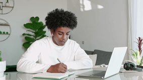 African American teenage guy student using computer watching webinar, learning english online virtual elearning class, taking professional web course writing notes and talking sitting at home table.