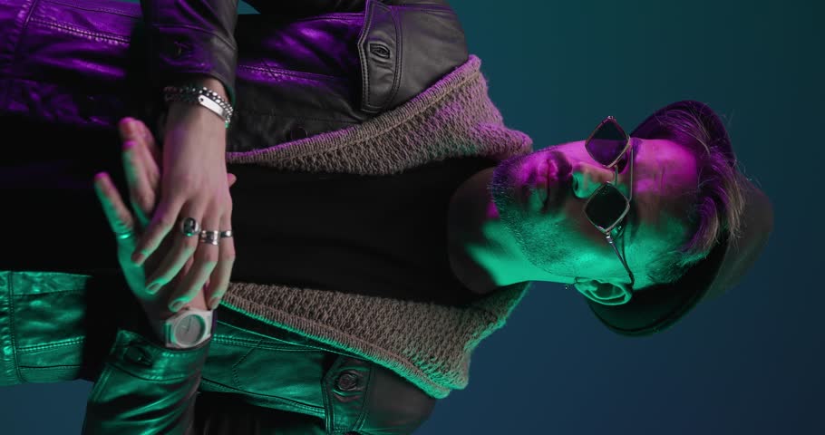 close up video of casual young man with cool glasses rubbing palms, adjusting jacket and looking to side on a colorful vertical video