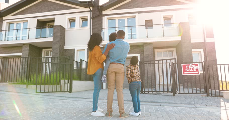 Back view on happy African American family with small children standing at new house at suburb and looking on it. Rear on cheerful parents and kids moving in new home. Settlement in outskirt. Sold. Royalty-Free Stock Footage #1099562555