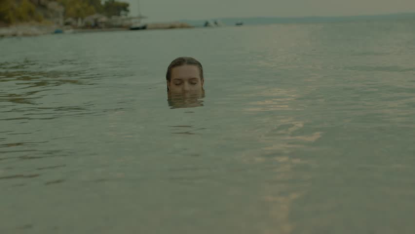 Portrait of a young girl in a swimsuit swims in the sea and smiles at sunset. Woman resting on vacation, enjoying time on the beach. | Shutterstock HD Video #1099563337
