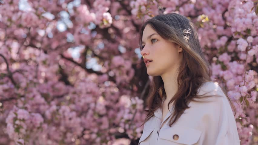 Sick caucasian woman using white napkins during runny nose outdoors. Side view of brunette standing near blooming sakura tree and suffering from seasonal allergy. | Shutterstock HD Video #1099570225