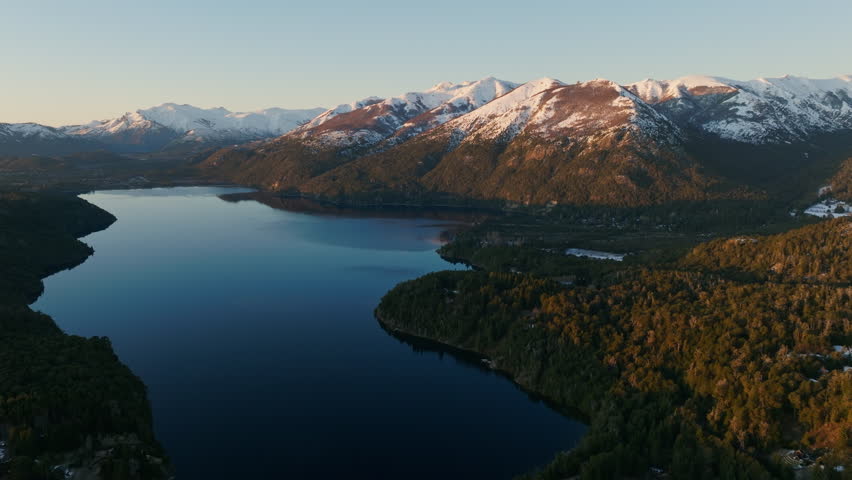 A wide drone flight panning from right to left, over the Nahuel Huapi lake with a view of the sunlit mountain range and lush green forest at Bariloche, Argentina during sunrise