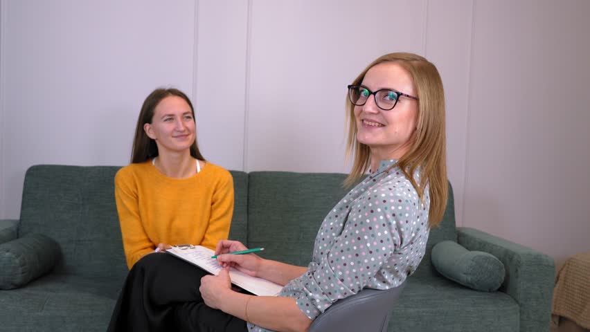 Two girls talking and discussing project. One of them turns to camera looking and smiling. Business project, work in team or in company. business women chatting in office sitting on chair and sofa. | Shutterstock HD Video #1099580733