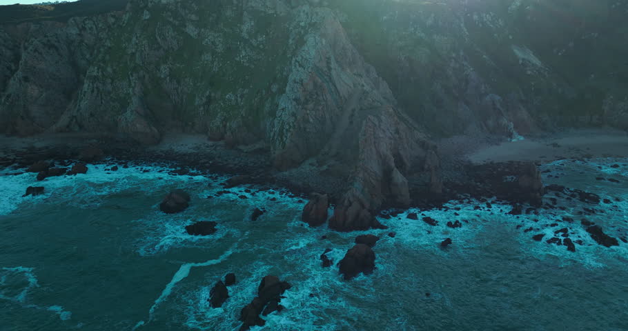 Aerial view of ocean blue waves breaking on a rocky mountain. Drone flies over the cost of Atlantic ocean against the morning sun.  | Shutterstock HD Video #1099581013