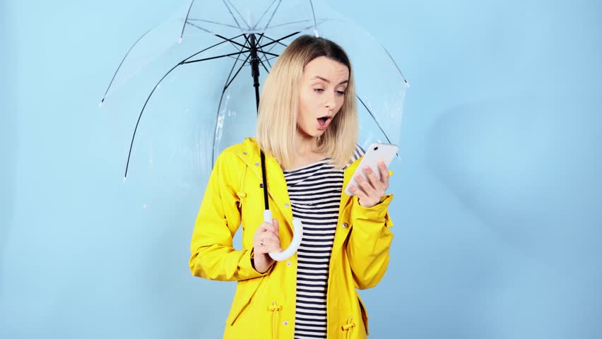 Boring woman in yellow raincoat scrolling products in internet store on smartphone under umbrella while find incredible unique offer product promotion wow surprised face expression on blue background | Shutterstock HD Video #1099581465