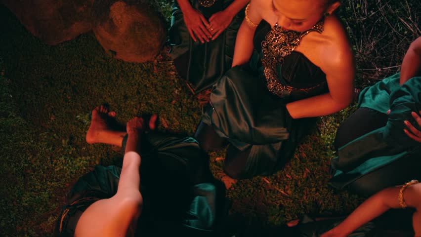 A high-angle shot of women in traditional costumes sitting on the grass at night. Their hands touch and arrange green silk cloth in a mysterious ritual.