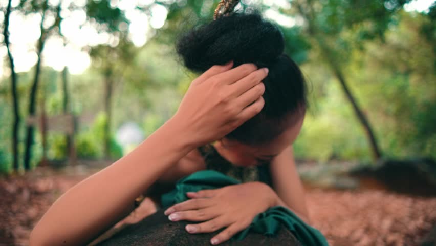 Close-up of a beautiful Indonesian woman in traditional costume and makeup. She adjusts her hair bun, then looks up, staring seriously at the camera in a forest setting.