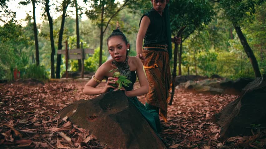 Low-angle shot of a man in traditional Javanese costume walking barefoot past a woman crouching by a rock. Mysterious narrative scene in a forest.