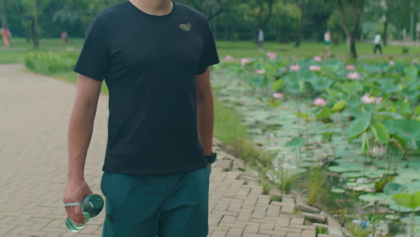 Tilt up shot of Caucasian man in sportswear walking in park and drinking water from bottle before morning run