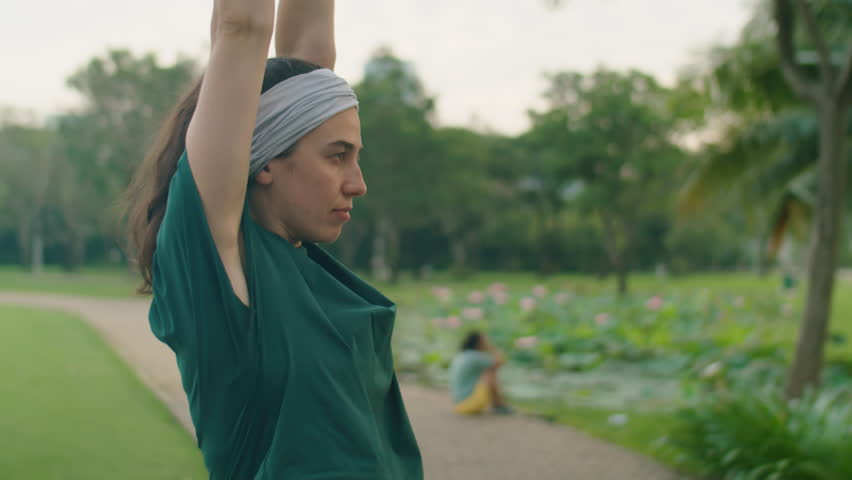 Medium arc shot of young woman in sportswear stretching arms, wrists and shoulders while doing warmup before training in park