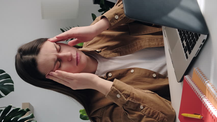 Vertical shot of tired young woman massaging temples, suffering from headache after computer work, sitting at table with laptop in room. Exhausted girl touching head, relieving pain, migraine concept | Shutterstock HD Video #1099585915