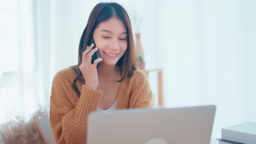 Happy young asian woman talking to client on mobile phone while using laptop discuss deal at home, Female customer make mobile call confirming online website shopping order delivery concept | Shutterstock HD Video #1099586399