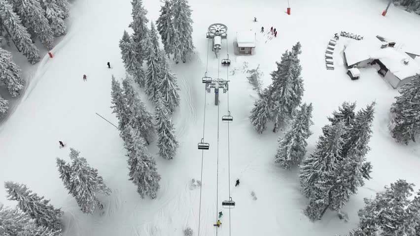 Aerial view over snowy mountain winter forest with chair lift at ski resort. Ski lift cable car top down drone shot. Winter landscape scene with snow covered trees. Outdoor tourism skiing snowboarding