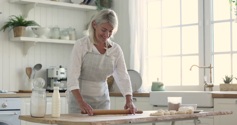 Beautiful mature woman wear apron cooking alone standing in kitchen, flattening dough for pastries, smile looking at camera enjoy cookery process on weekend at home. Chores, housework, homemade baking | Shutterstock HD Video #1099588923