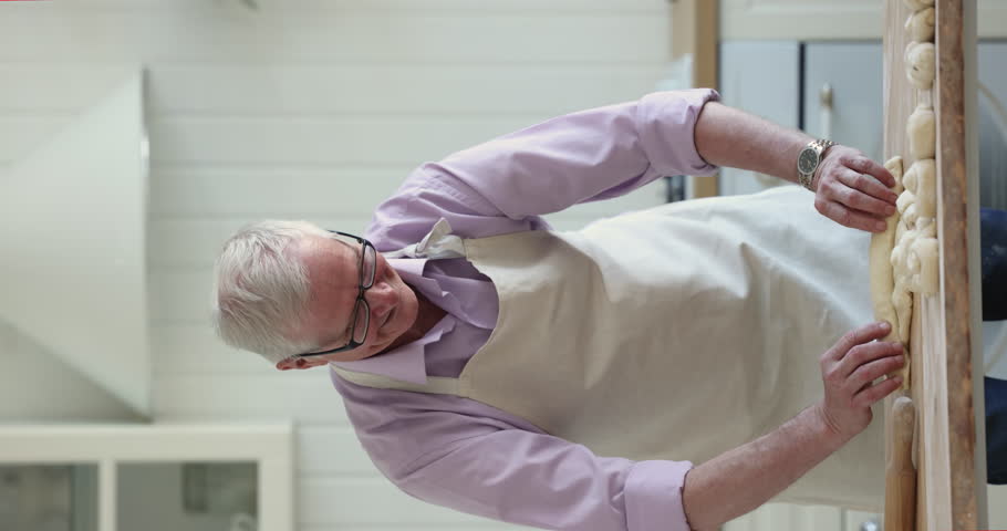 Senior gray-haired man wear apron, kneading homemade dough, prepare pastries, surprise for family or guests, enjoy cooking process standing alone in cozy domestic kitchen. Culinary hobby on retirement | Shutterstock HD Video #1099588925